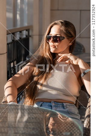 Woman Sunglasses Casual Summer Outfit Portrait. A woman with long brown hair sits in a chair, wearing sunglasses and a white tank top. She rests her hand on her chin and looks off to the side. 117352924