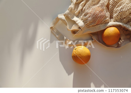 String bag with oranges fruit on beige table background in sunlight. Long shadows. Moody summer vacation concept. Healthy fresh food, lifestyle web banner, decorative frame. Top, flat lay. Copy space. 117353074