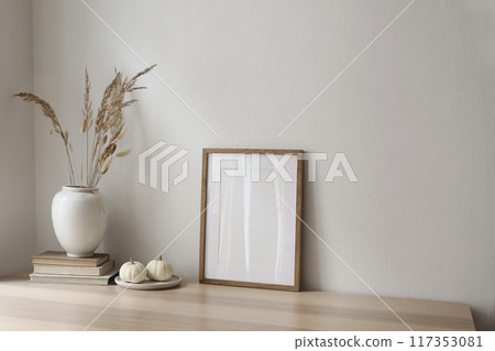 Elegant neutral poster mockup. Wooden picture frame on beige table. Vase with dry grass and old books and little white pumpkins. Empty wall background. Scandinavian interior, home. Autumn decor. 117353081