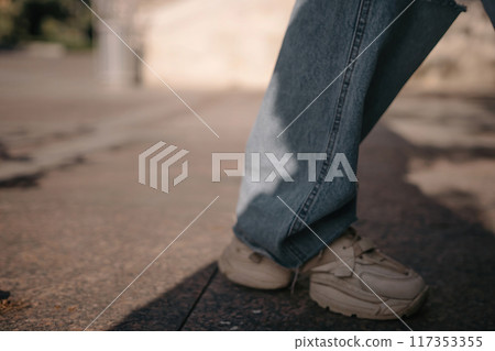 A woman is walking down a sidewalk wearing ripped jeans and a gray jacket. The image conveys a casual and relaxed atmosphere, as the woman is enjoying her walk. A woman is walking down a sidewalk wearing ripped jeans and a gray jacket. The image conveys a casual and relaxed atmosphere, as the woman is enjoying her walk. 117353355