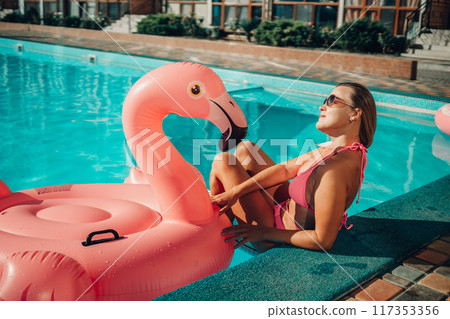 A woman in a pink bikini is sitting on a pink inflatable flamingo in a pool. The scene is playful and fun, with the woman enjoying her time in the water. A woman in a pink bikini is sitting on a pink inflatable flamingo in a pool. The scene is playful and fun, with the woman enjoying her time in the water. 117353356