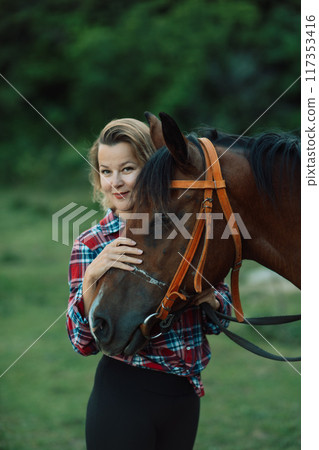 Happy blonde with horse in forest. Woman and a horse walking through the field during the day. Dressed in a plaid shirt and black leggings. 117353416
