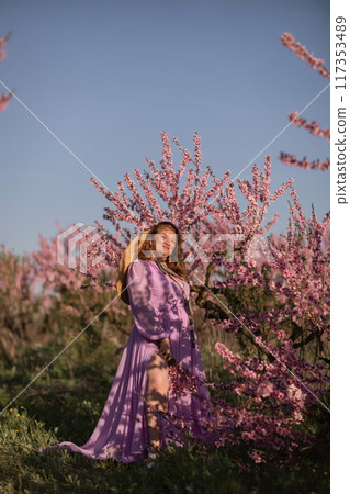 Woman blooming peach orchard. Against the backdrop of a picturesque peach orchard, a woman in a long pink dress and hat enjoys a peaceful walk in the park, surrounded by the beauty of nature. Woman blooming peach orchard. Against the backdrop of a picturesque peach orchard, a woman in a long pink dress and hat enjoys a peaceful walk in the park, surrounded by the beauty of nature. 117353489