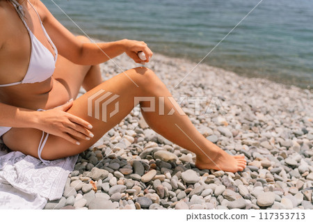 A woman is sitting on a beach with her legs crossed and a bottle of sunscreen in her hand. A woman is sitting on a beach with her legs crossed and a bottle of sunscreen in her hand. 117353713