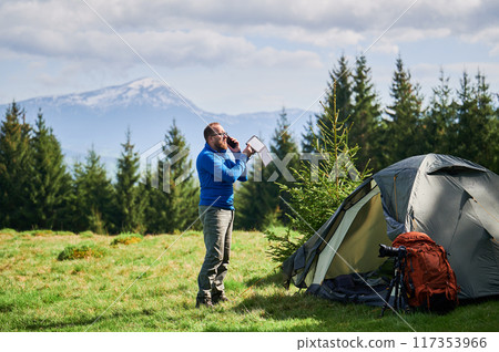 Man tourist stands beside tent, talking on phone and holding portable solar panel. Orange backpack and camera tripod nearby. Snow-capped mountains and lush evergreen trees under partly cloudy sky. Man tourist stands beside tent, talking on phone and holding portable solar panel. Orange backpack and camera tripod nearby. Snow-capped mountains and lush evergreen trees under partly cloudy sky. 117353966