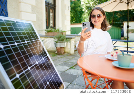 Young woman charging her smartphone from solar panel while relaxing. Pretty girl drinking coffee and checking her phone next to solar battery. Conscious female choosing sustainable lifestyle. 117353968