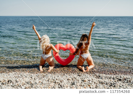 Two women are posing on a beach with a red heart-shaped inflatable raft 117354268