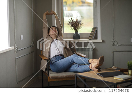 Smiling Young caucasian Female Manager Sitting On Chair Stretching Her Arms. Work-life-balance Smiling Young caucasian Female Manager Sitting On Chair Stretching Her Arms. Work-life-balance 117354678