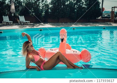 A woman is laying on a pink inflatable flamingo in a pool. The scene is bright and cheerful, with the pink flamingo adding a playful touch to the image. A woman is laying on a pink inflatable flamingo in a pool. The scene is bright and cheerful, with the pink flamingo adding a playful touch to the image. 117355617