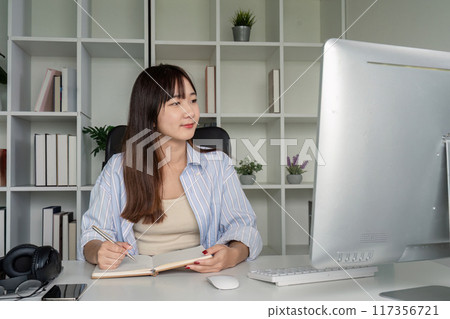 Young Student Studying Online at Home Using Desktop Computer in Modern Home Office Setting with Books and Plants 117356721