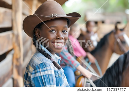 Joyful young girl with a wide smile wearing a cowboy hat while riding a horse, with other children in the background 117357757