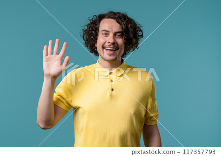 Friendly young man waving hand - hello, or goodbye, chao, salute. Parting, say bye to camera. Handsome man on blue studio background. Friendly young man waving hand - hello, or goodbye, chao, salute. Parting, say bye to camera. Handsome man on blue studio background. 117357793