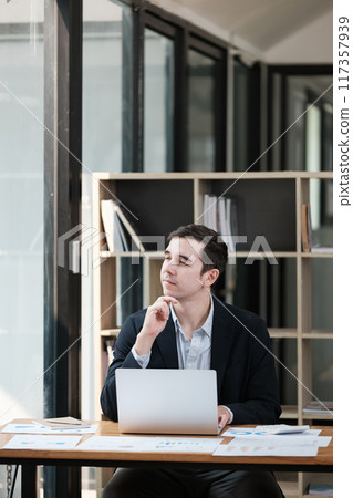 A man is sitting at a desk with a laptop and a stack of papers A man is sitting at a desk with a laptop and a stack of papers 117357939
