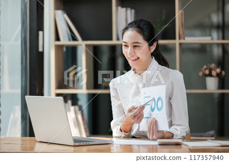 A woman is sitting at a desk with a laptop and a piece of paper A woman is sitting at a desk with a laptop and a piece of paper 117357940