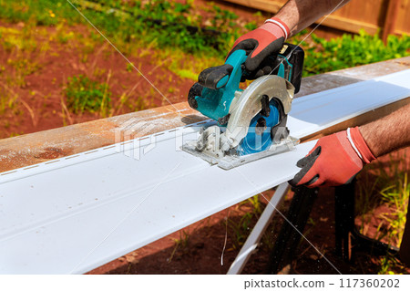 Working with circular handsaw, worker cuts plastic vinyl siding to size before it is installed 117360202