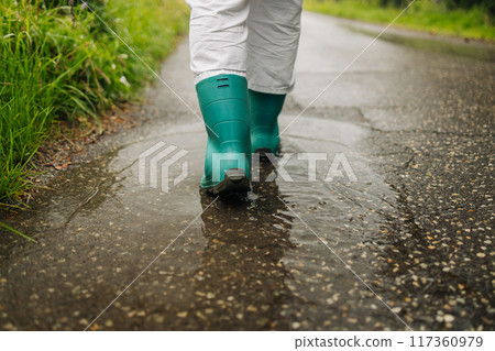 Woman wearing rain rubber boots walking running and jumping into puddle with water splash and drops in autumn rain. 117360979