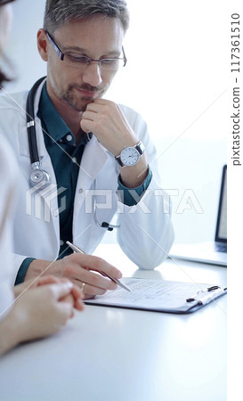 Doctor and a patient. The physician, wearing a white medical coat over a green shirt, gesturing with his hands during a consultation in the clinic. Medicine concept Doctor and a patient. The physician, wearing a white medical coat over a green shirt, gesturing with his hands during a consultation in the clinic. Medicine concept 117361510
