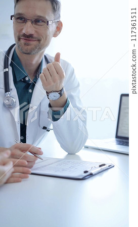 Doctor and a patient. The physician, wearing a white medical coat over a green shirt, gesturing with his hands during a consultation in the clinic. Medicine concept Doctor and a patient. The physician, wearing a white medical coat over a green shirt, gesturing with his hands during a consultation in the clinic. Medicine concept 117361511
