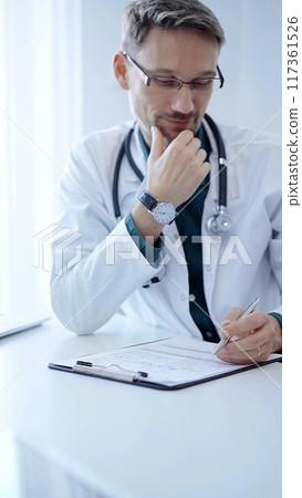 Doctor and a patient. The physician, wearing a white medical coat over a green shirt, gesturing with his hands during a consultation in the clinic. Medicine concept 117361526