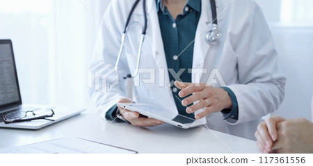Doctor and a patient. The physician, wearing a white medical coat over a green shirt, is using tablet computer during a consultation in the clinic. Medicine concept Doctor and a patient. The physician, wearing a white medical coat over a green shirt, is using tablet computer during a consultation in the clinic. Medicine concept 117361556