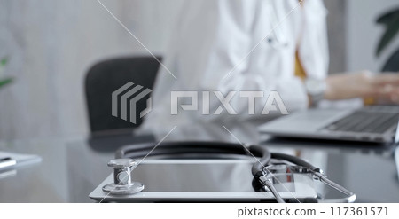Close-up of doctor's desk with stethoscope and tablet computer. Physician is using a laptop at the background. Medicine concept 117361571