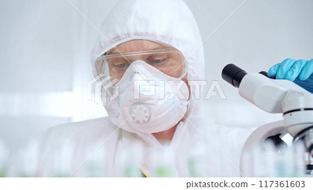 Professional scientist in protective gear is using microscope in a laboratory setting and alarm red light. Science and medicine concepts 117361603