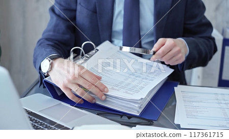 Adult businessman or auditor examining documents with magnifying glass. Professional in a suit scrutinizes financial reports, detail-focused work concept Adult businessman or auditor examining documents with magnifying glass. Professional in a suit scrutinizes financial reports, detail-focused work concept 117361715