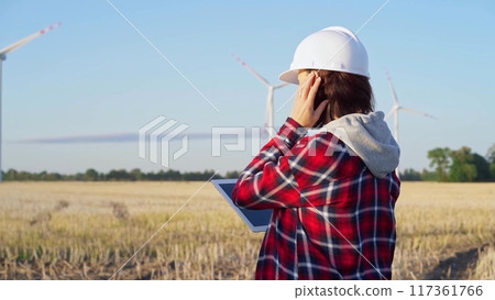 Female engineer wearing a white protective helmet is taking notes with a tablet computer on a field with wind turbines, as the sun sets. Clean energy and engineering 117361766