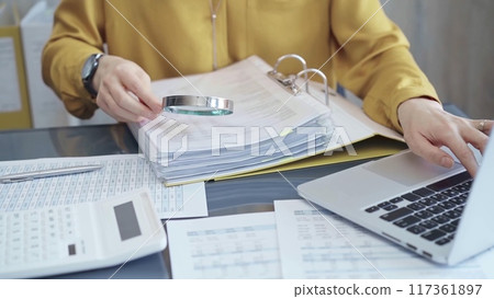 Female auditor in business attire inspecting financial reports with magnifying glass, surrounded by documents and a laptop and calculator. Business audit and taxes concept 117361897