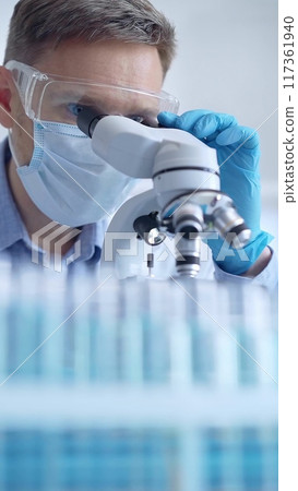 Man scientist with protective glasses, mask and gloves is using microscope in laboratory, vertical portrait of researcher. Microbiology and science Man scientist with protective glasses, mask and gloves is using microscope in laboratory, vertical portrait of researcher. Microbiology and science 117361940