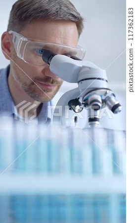Man scientist with protective glasses and gloves is using microscope in laboratory, vertical portrait of researcher. Microbiology science concept Man scientist with protective glasses and gloves is using microscope in laboratory, vertical portrait of researcher. Microbiology science concept 117362183