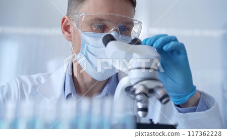 Man scientist with protective glasses, mask and gloves is using microscope in laboratory, portrait of researcher. Microbiology science concept 117362228