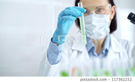 Woman scientist with protective gloves and glasses examining lab tube with a green fluid. Microbiology and science concept 117362252