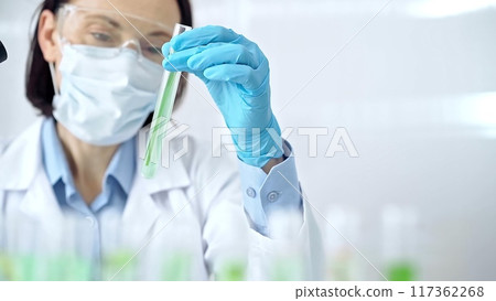 Woman scientist with protective gloves and glasses examining lab tube with a green fluid. Microbiology and science concept Woman scientist with protective gloves and glasses examining lab tube with a green fluid. Microbiology and science concept 117362268