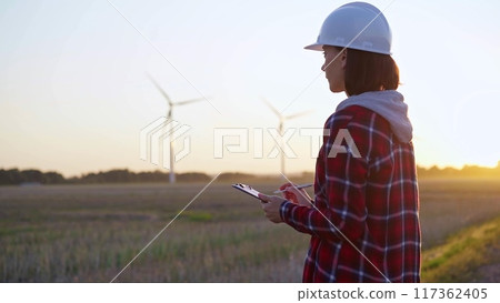 Adult woman engineer wearing white cask is taking notes on a clipboard on a field with wind turbines Adult woman engineer wearing white cask is taking notes on a clipboard on a field with wind turbines 117362405