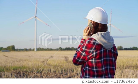 Woman engineer is using a smartphone in a field with wind turbines as the sun sets. Concept of clean energy and engineering audit 117362450