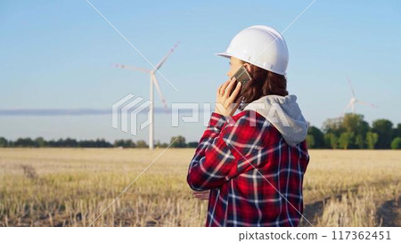 Woman engineer is using a smartphone in a field with wind turbines as the sun sets. Concept of clean energy and engineering audit 117362451