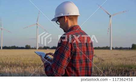 Man engineer at work is taking notes with a tablet computer in a field with wind turbines, as the sun sets. Clean energy concept Man engineer at work is taking notes with a tablet computer in a field with wind turbines, as the sun sets. Clean energy concept 117362458
