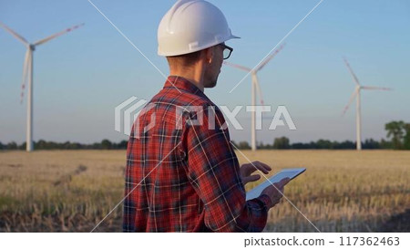Man engineer at work is wearing a white protective helmet and taking notes with a tablet computer in a field with wind turbines, as the sun sets. Clean energy concept Man engineer at work is wearing a white protective helmet and taking notes with a tablet computer in a field with wind turbines, as the sun sets. Clean energy concept 117362463
