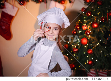 Cheerful girl in chef's hat shows looking through baking pan of cookies on Christmas background Cheerful girl in chef's hat shows looking through baking pan of cookies on Christmas background 117364194