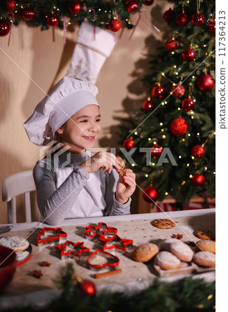 Cheerful girl in chef's hat eating homemade cookies on Christmas background 117364213