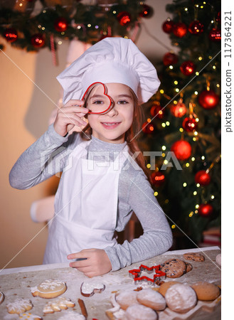 Cheerful girl in chef's hat shows looking through baking pan of cookies on Christmas background 117364221