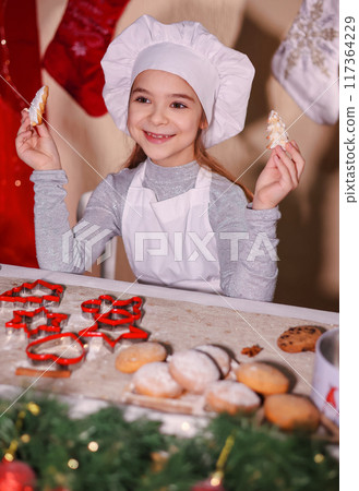 A cheerful girl in a chef's hat shows Christmas gingerbread cookies in the shape of a Christmas tree. 117364229