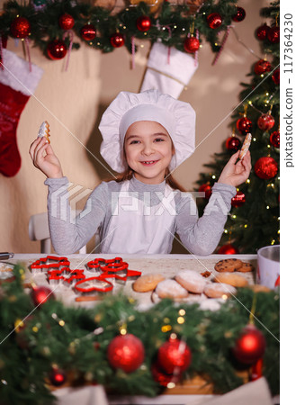A girl in a chef's hat smiles and shows Christmas gingerbread cookies in the shape of a Christmas tree. A girl in a chef's hat smiles and shows Christmas gingerbread cookies in the shape of a Christmas tree. 117364230