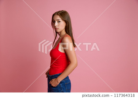 Side view portrait of young girl in red top and jeans looking at camera with serious expression against pink studio background Side view portrait of young girl in red top and jeans looking at camera with serious expression against pink studio background 117364318