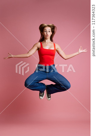 Full-length image of young beautiful girl in jeans and red top jumping in yoga pose against pink studio background. Meditation and energy 117364323