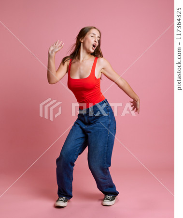 Hip-hop dance class. Portrait of young beautiful girl in casual clothes, red top and jeans cheerfully posing, dancing against pink studio background 117364325