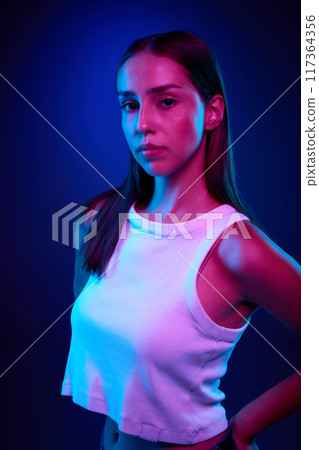 Portrait of beautiful young woman in casual clothes, white top posing with calm face against gradient blue studio background in neon light. Confident look Portrait of beautiful young woman in casual clothes, white top posing with calm face against gradient blue studio background in neon light. Confident look 117364356