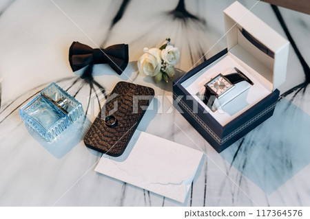 Groom's wedding essentials on table, featuring rings on phone, bow tie, boutonniere, perfume, belt 117364576