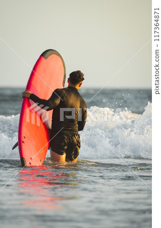 Surfer in wetsuit getting into the sea with his red surfboard Surfer in wetsuit getting into the sea with his red surfboard 117364871